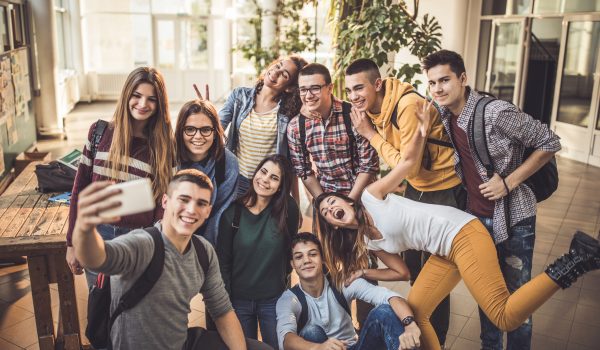 Large group of happy high school students having fun while taking a selfie with mobile phone in a hallway.